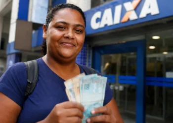 Mulher segura notas de dinheiro em frente a uma agência bancária, representando o pagamento do 4º lote da restituição do IRPF 2026.