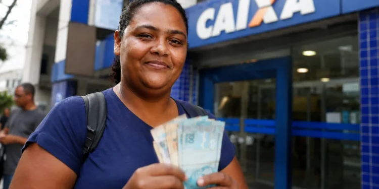 Mulher segura notas de dinheiro em frente a uma agência bancária, representando o pagamento do 4º lote da restituição do IRPF 2026.