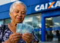 Idosa segura cédulas de real em frente a uma agência da Caixa, representando os pagamentos do INSS de até um salário mínimo em agosto.