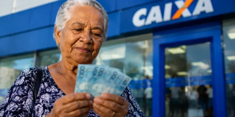 Idosa segura cédulas de real em frente a uma agência da Caixa, representando os pagamentos do INSS de até um salário mínimo em agosto.