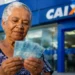 Idosa segura cédulas de real em frente a uma agência da Caixa, representando os pagamentos do INSS de até um salário mínimo em agosto.