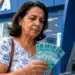 Beneficiária do INSS com notas de dinheiro em frente a um caixa eletrônico da Caixa, representando os pagamentos de agosto para segurados que recebem acima do salário mínimo.