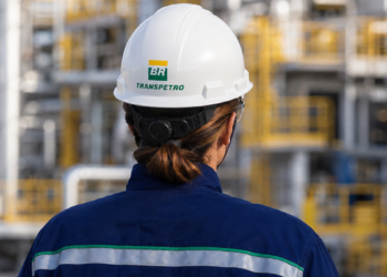 Profissional uniformizada da Transpetro vista de costas em área industrial, com capacete de segurança identificado com a marca da empresa.