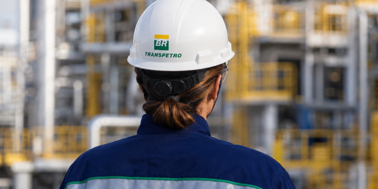 Profissional uniformizada da Transpetro vista de costas em área industrial, com capacete de segurança identificado com a marca da empresa.