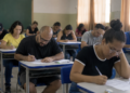 Candidatos realizando prova de concurso público em sala de aula com questões objetivas e discursivas.