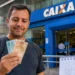 Homem segurando notas de real em frente a uma agência da Caixa, representando o pagamento do quarto lote da restituição do IRPF 2026.