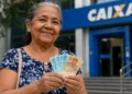 Idosa segura cédulas em frente a uma agência da Caixa, em referência ao reajuste do salário mínimo e aos benefícios do INSS e BPC em 2027.