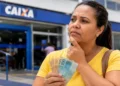 Mulher segura notas de real em frente a uma agência da Caixa, representando os rumores sobre um possível valor de R$ 1.200 para o Bolsa Família em 2027.
