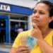Mulher segura notas de real em frente a uma agência da Caixa, representando os rumores sobre um possível valor de R$ 1.200 para o Bolsa Família em 2027.
