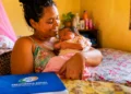 Mãe segura recém-nascido em casa, com documento da Previdência Social sobre a mesa, representando o salário-maternidade do INSS.