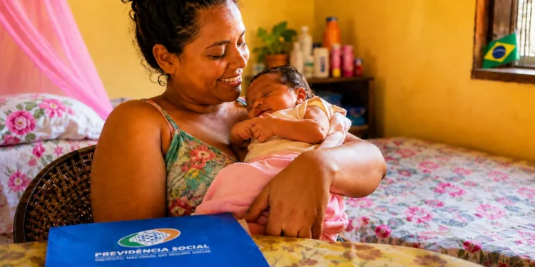 Mãe segura recém-nascido em casa, com documento da Previdência Social sobre a mesa, representando o salário-maternidade do INSS.