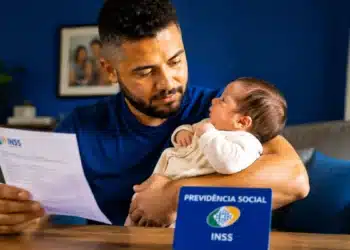 Pai segura bebê recém-nascido enquanto consulta documento relacionado ao INSS sobre salário-maternidade.