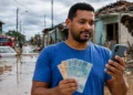 Pessoa consulta o celular e segura dinheiro em área atingida por enchente durante situação de calamidade relacionada ao Saque Calamidade do FGTS.