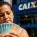 Mulher em frente a uma agência da Caixa segurando cédulas de real, representando o Saque Calamidade do FGTS liberado para moradores do Rio Grande do Sul.