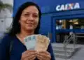 Mulher segura cédulas em frente a uma agência da Caixa, representando a liberação do saque do FGTS de até R$ 6,2 mil para moradores de 19 cidades.
