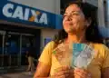 Mulher segurando notas de real em frente a uma agência da Caixa para representar o saque do FGTS de até R$ 6.220 destinado a moradores de RS, MG e SP
