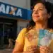 Mulher segurando notas de real em frente a uma agência da Caixa para representar o saque do FGTS de até R$ 6.220 destinado a moradores de RS, MG e SP