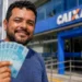 Trabalhador segura cédulas diante de uma agência da Caixa, ilustrando o saque de até R$ 6,2 mil do FGTS.