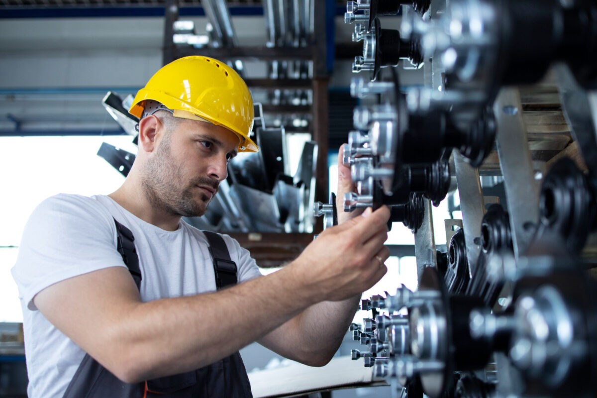 Imagem ilustrativa de profissional de capacete amarelo e macacão ajustando componentes metálicos em uma estrutura industrial, em referência à atuação do técnico em instrumentação.