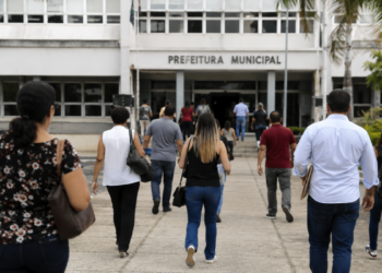 Pessoas caminham em direção a uma prefeitura municipal durante período de inscrições para concursos públicos.