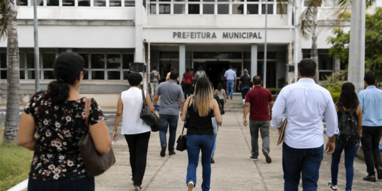 Pessoas caminham em direção a uma prefeitura municipal durante período de inscrições para concursos públicos.