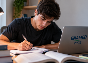 Jovem escrevendo em caderno ao lado de laptop com logo ENAMED 2026 em mesa iluminada.
