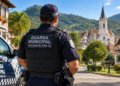 Guarda Municipal de costas, ao lado de viatura em rua arborizada com igreja ao fundo.