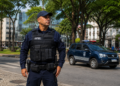 Guarda Municipal de uniforme tático observa rua com viatura oficial ao fundo em centro de cidade arborizada.