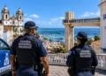 Dois agentes da Guarda Municipal de Salvador observam o Elevador Lacerda e o mar em dia ensolarado.