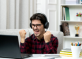 Jovem de camisa xadrez e óculos celebra de fones diante do notebook com punhos erguidos em ambiente doméstico.