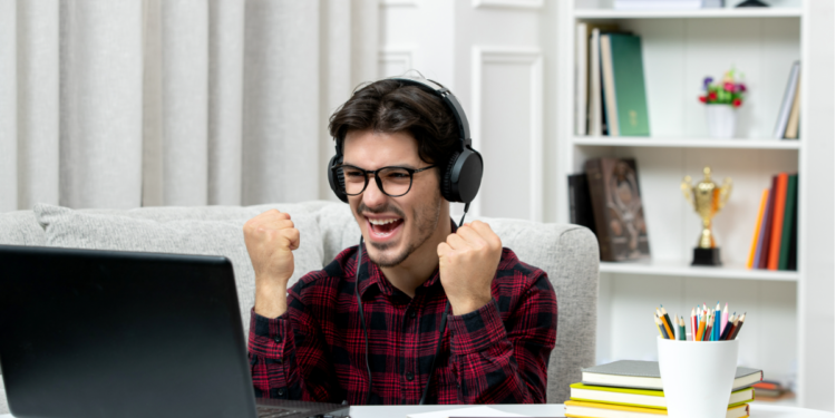 Jovem de camisa xadrez e óculos celebra de fones diante do notebook com punhos erguidos em ambiente doméstico.