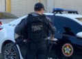 Policial civil de colete armado em frente a viatura preta e branca.