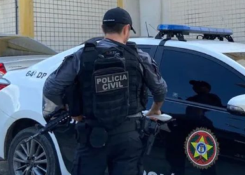 Policial civil de colete armado em frente a viatura preta e branca.