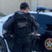 Policial civil de colete armado em frente a viatura preta e branca.