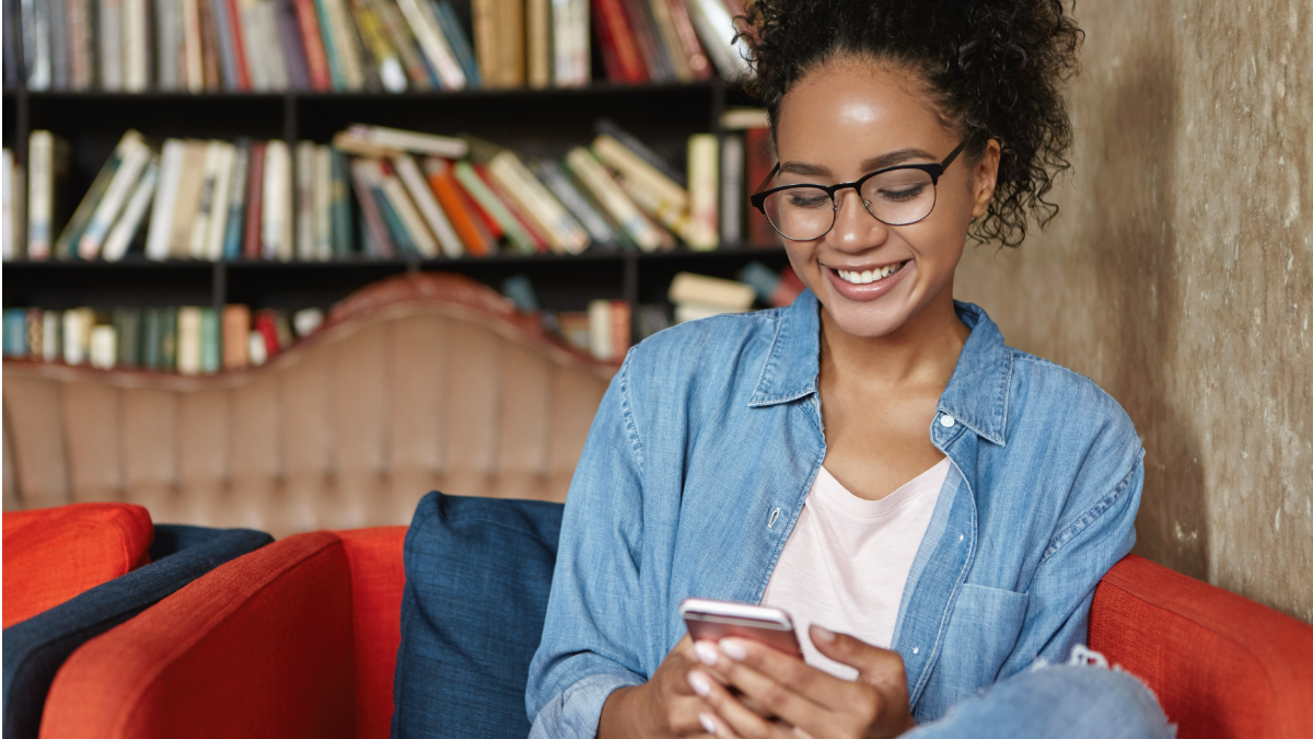 Jovem sorrindo e mexendo no celular em biblioteca.