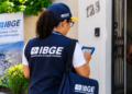 Recenseadora do IBGE, com colete azul, entrevista moradora na porta de casa, com banner do IBGE ao fundo.