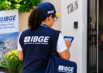 Recenseadora do IBGE, com colete azul, entrevista moradora na porta de casa, com banner do IBGE ao fundo.