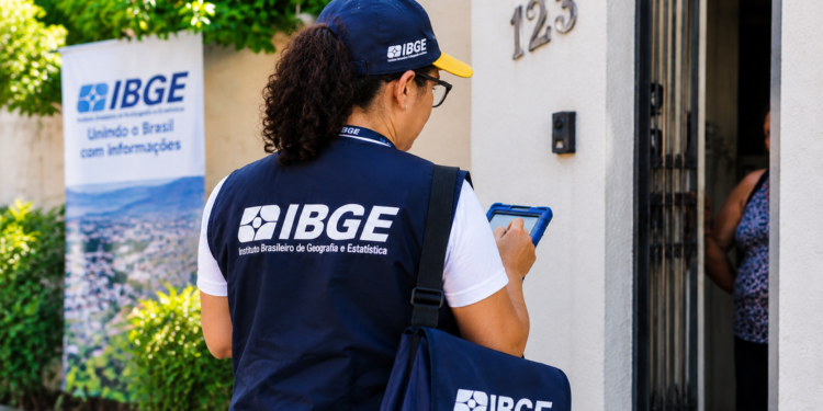 Recenseadora do IBGE, com colete azul, entrevista moradora na porta de casa, com banner do IBGE ao fundo.