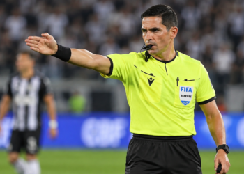Árbitro masculino com uniforme amarelo da FIFA 2024 apita e aponta braço direito durante jogo.