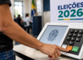 Eleitor pressionando botão na urna eletrônica diante de cartaz "Eleições 2026" e bandeira do Brasil.