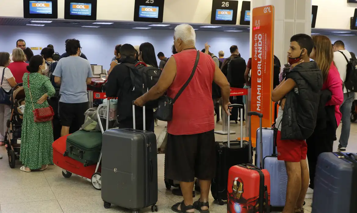 Passageiros com malas aguardando atendimento em fila de balcão de check-in em um terminal de aeroporto.