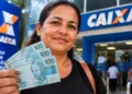 Mulher segura notas de real em frente a uma agência da Caixa, representando a antecipação do pagamento do Bolsa Família de setembro.