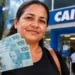Mulher segura notas de real em frente a uma agência da Caixa, representando a antecipação do pagamento do Bolsa Família de setembro.
