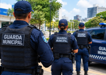 Três agentes da Guarda Municipal de colete e boné patrulham rua ao lado de viatura marcada.