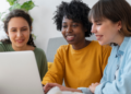 Três pessoas analisam informações sobre concursos públicos em um notebook.