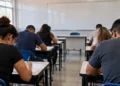 Candidatos realizando prova de concurso público em sala de aula com folhas de questões sobre as mesas.