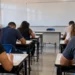 Candidatos realizando prova de concurso público em sala de aula com folhas de questões sobre as mesas.