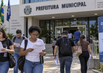 Pessoas se preparando para concurso público em frente a uma prefeitura municipal com documentos de inscrição em mãos.