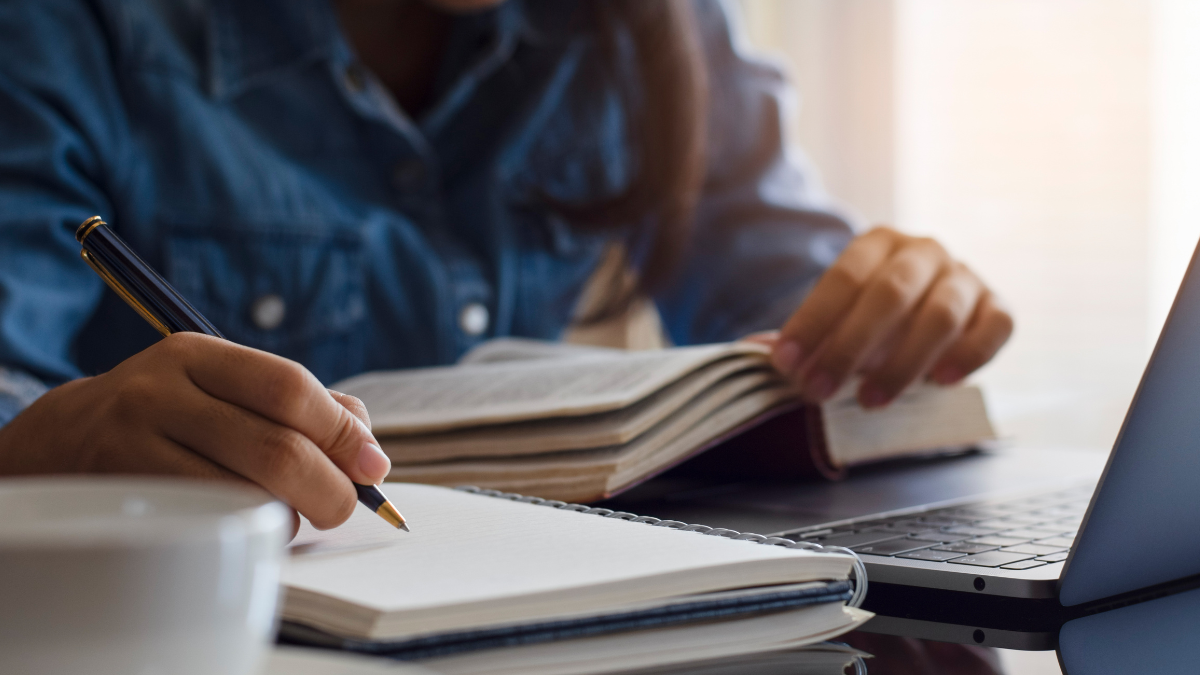 Pessoa estudando para concursos públicos com livros, caderno e notebook sobre a mesa.