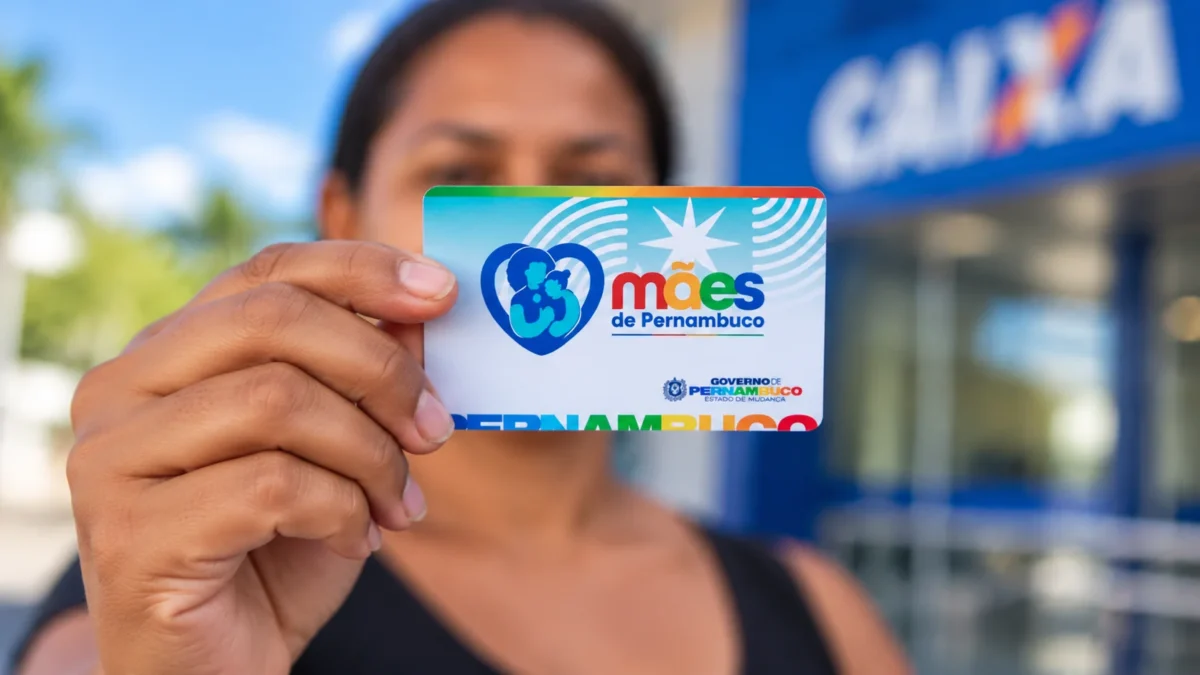 Cartão do programa Mães de Pernambuco segurado por beneficiária em frente a uma agência da Caixa